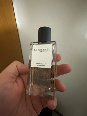 Le Persona Whispering Kiss parfym - Le Persona Whispering Kiss extrait de parfum i en stilren, rektangulär glasflaska med svart lock. Flaskan har en transparent design som visar den bärnstensfärgade vätskan inuti och en elegant etikett framtill. Perfekt för dig som gillar exklusiva dofter. OBS! Saknar förpackning!