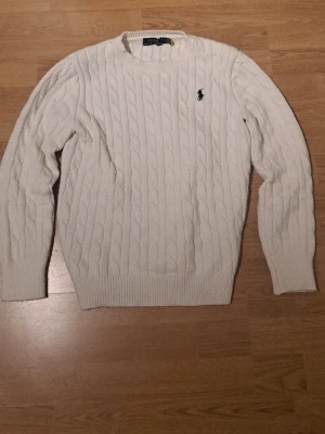 Vit kabelstickad tröja från Polo Ralph Lauren - Kabelstickad vit tröja från Polo Ralph Lauren med klassisk svart broderad logga på bröstet. Tröjan har rund halsringning, ribbade muddar och nederkant. Perfekt för dig som gillar stilrena och tidlösa plagg.