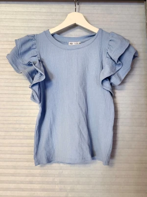 Ljusblå topp med volangärmar från Zara - Supersöt ljusblå topp från Zara med ribbad struktur och volangärmar. Rund halsringning och mjukt, stretchigt material som sitter bekvämt. Perfekt för dig som gillar en feminin och trendig stil.
