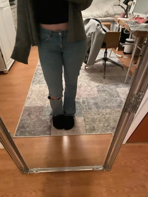 Ljusblå jeans  - Säljer ett par ljusblå bootcut jeans med klassisk femficksdesign och ett snyggt hål på ena knät. Jeansen har låg midja och stängs med knapp och dragkedja. Perfekta för en avslappnad och trendig look.
