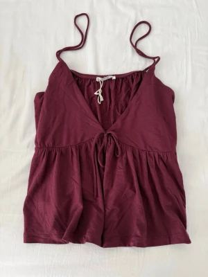 Vinröd topp med tunna axelband - Snygg vinröd topp med tunna axelband och knytning framtill. Djup V-ringning och lätt rynkad nedtill. Storleken är M, 14+❣️❣️aldrig använd.