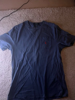 Blå t-shirt från Polo Ralph Lauren - Snygg blå t-shirt från Polo Ralph Lauren med klassisk passform. T-shirten har en bröstficka med den ikoniska röda loggan broderad på fickan. Tillverkad i mjuk bomull och har korta ärmar. Perfekt till jeans eller shorts.