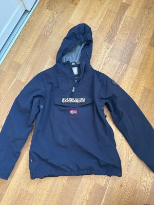 Mörkblå fodrad anorak/jacka från Napapijri - Snygg mörkblå vindjacka från Napapijri med huva och halv dragkedja. Stor ficka framtill med logotyp och norsk flagga. Jackan har elastisk nederkant och är tillverkad i slitstarkt material, perfekt för blåsiga dagar.