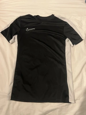 Svart Nike sporttröja med vita detaljer - Snygg svart sporttröja från Nike med vita sidopaneler och vit logga på bröstet. Tröjan har korta ärmar och är tillverkad i ett lätt, snabbtorkande funktionsmaterial som passar perfekt till träning eller match.