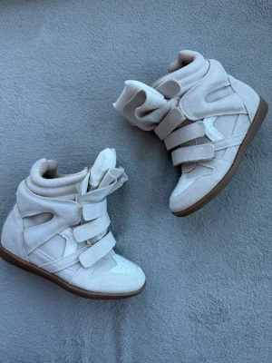 Vita höga sneakers med kardborreband - Stilrena vita sneakers med högt skaft och flera breda kardborreband. Skorna har en chunky design, ovandel i mockaliknande material och detaljer i vitt läder. Yttersulan är i gummi med bra grepp. Perfekta för dig som gillar statement-sneakers med streetkänsla.