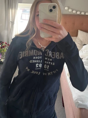 Mörkblå hoodie från Abercrombie & Fitch - Säljer en mörkblå hoodie från Abercrombie Fitch med vit text på bröstet och dragkedja framtill. Hoodien har huva och två fickor. Storlek M men liten passform