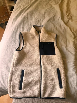 Beige fleeceväst från Oceanswe - Mysig beige fleeceväst från Oceanswe med svarta detaljer. Västen har hög krage, dragkedja framtill, två fickor med svarta kanter och en svart bröstficka med dragkedja. Perfekt för lager-på-lager och chill stil.