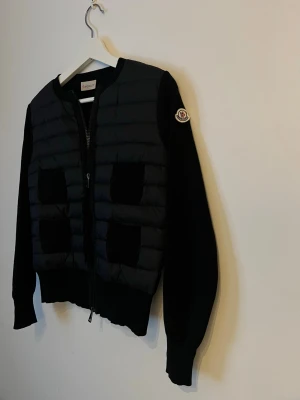 Svart cardiganjacka moncler  - Snygg svart cardiganjacka från Moncler med quiltad front och stickade ärmar. Jackan har dragkedja framtill, ribbade muddar och fyra dekorativa fickor på framsidan. Moncler-logga på ena ärmen. Perfekt för lager på lager och streetstyle.