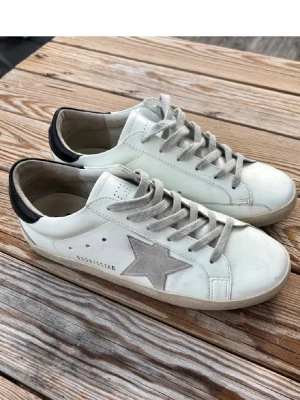 Golden Goose Superstar vita sneakers - Golden Goose Superstar sneakers i vitt läder med grå mockastjärna på sidan, grå skosnören och svart detalj på hälen. Klassisk låg modell med vintage-inspirerad look och perforerade detaljer vid snörningen. Perfekta för en avslappnad och trendig stil.