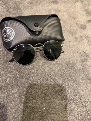 Ray-Ban svarta solglasögon - Säljer ett par klassiska Ray-Ban solglasögon med svart fodral. Fodralet har en snygg struktur och Ray-Ban-logga på sidan. Perfekt för dig som vill ha en ikonisk och stilren accessoar till sommaren.