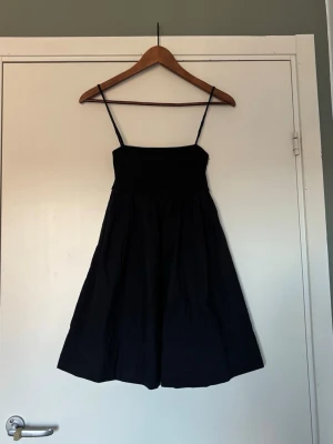 Svart kort klänning med smala axelband - En svart, kort klänning med smala axelband och rynkad överdel. Klänningen har en lätt utställd kjol och är tillverkad i ett mjukt tyg 