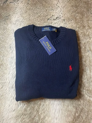 Mörkblå stickad tröja Polo Ralph Lauren - Mörkblå stickad tröja från Polo Ralph Lauren med klassisk rund halsringning och ribbade muddar. Tröjan har den ikoniska röda Polo-loggan broderad på bröstet. Perfekt för en stilren och avslappnad look.