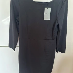 Svart kortklänning från Icona - Snygg svart klänning från Icona med enkel och stilren design. Modellen är figurnära med trekvartsärm och rund halsringning. Klänningen har en diskret söm i midjan och dragkedja bak. Perfekt för dig som gillar minimalistisk stil.