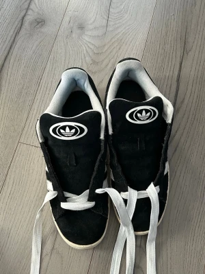 Svarta Adidas sneakers med vita ränder - Klassiska svarta Adidas campus sneakers med vita ränder på sidorna och vit Adidas-logga på plösen. Skorna har vit sula och gummiyttersula i brunt. Ovandelen är i mocka och de har vita skosnören. Perfekt för en stilren look.