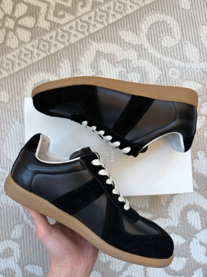 Maison Margela Gats - Säljer ett par helt nya Maison Margela GAT i svart färg med gumsoles. En ikonisk och tidlös sneaker som fungerar till både uppklätt och casual.  Tillverkade i svart läder med matchande mockadetaljer och gummifärgad sula för en stilren, minimalistisk look. Klassisk låg silhuett med snörning framtill.  Skick: Helt nya Färg: Svart med gummi sula Modell: GAT  Storlek: 42 Hör av dig vid frågor 