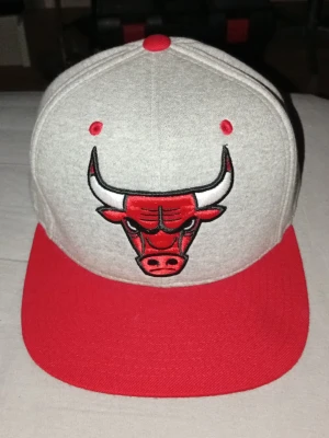 Chicago Bulls keps grå/röd - Snygg keps med Chicago Bulls-logga broderad framtill. Kepsen är ljusgrå med röd skärm, röda ventilationshål och knapp på toppen. Klassisk snapback-modell med platt skärm, perfekt för dig som gillar streetwear och sportig stil.