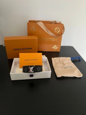 Louis Vuitton svart armband Monogram - Snyggt svart armband från Louis Vuitton med klassiskt monogrammönster och silverfärgad LV-logga. Tillverkat i exklusivt skinn med detaljer i silver. Kommer med original ask, dustbag och påse. Perfekt accessoar för dig som gillar lyx och stil.