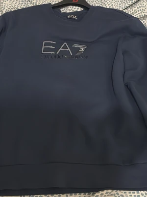 Mörkblå EA7 Emporio Armani hoodie - Snygg mörkblå hoodie från EA7 Emporio Armani med stort logotryck på bröstet. Klassisk modell med rund halsringning och mjukt material som känns skönt mot huden. Perfekt för en stilren och avslappnad look.