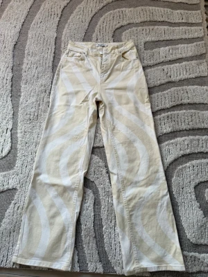 Beige och vita mönstrade jeans - Säljer ett par beige jeans med vita, vågiga mönster. Det är Misslisibells kollektion från NAKD. Byxorna har raka ben, klassisk femficksmodell och hög midja. Materialet är denim och de har en cool retro vibe som sticker ut i mängden. Använda 3-4 gånger. En liten fläck finns, skriv för en bättre bild på den. 