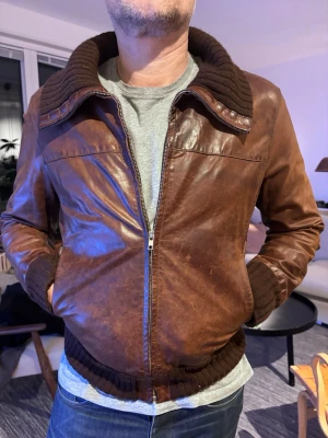 Brun skinnjacka med stickad krage - Snygg brun skinnjacka med dragkedja och bred, stickad krage samt muddar i brunt. Jackan har en klassisk modell med ribbade detaljer vid ärmslut och midja. Perfekt för dig som vill ha en tidlös och cool look.