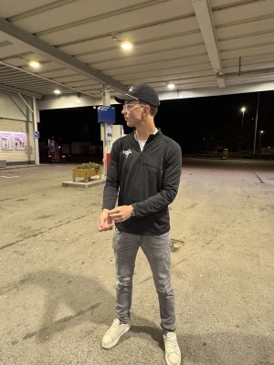 Golftröja - Svart långärmad tröja från Enclave med dragkedja vid halsen och vit logotyp på bröstet. Tröjan har en rak passform och är tillverkad i ett mjukt material som ger en avslappnad och stilren look. Perfekt för dig som gillar streetwear.