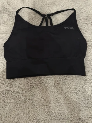 Svart sport-bh från FUZY med korsade band - Svart sport-bh från FUZY med bred resår nedtill och snygga korsade band i ryggen. Tillverkad i stretchigt funktionsmaterial som passar perfekt för träning. Enkel och stilren design med diskret logga framtill. Bh.n har uttagbara kuddar. Använd någon enstaka gång, köpt för ca 1 år sedan i en sportbutik. Använder inte då den är för stor. Texten där storleken står blev skadad vid tvätt, men annars har den inga tecken på användning. :))