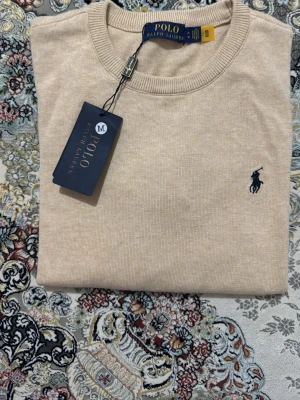 Beige stickad tröja Polo Ralph Lauren - Snygg beige stickad tröja från Polo Ralph Lauren med klassisk rund halsringning och den ikoniska mörkblå logotypen broderad på bröstet. Tröjan är tillverkad i mjukt material och har en stilren, enkel design som passar till det mesta.