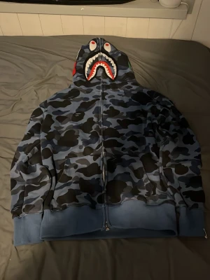 BAPE blå camo shark hoodie - Säljer en blå camo zip hoodie från BAPE med det ikoniska hajansiktet på huvan och broderade bokstäver på sidan. Hoodien har hel dragkedja, fickor fram och ribbade muddar. Materialet är mjuk bomullsmix och färgerna går i blått, svart och grått. Det står M men den sitter som S