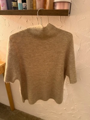 Beige stickad tröja med kort ärm - Mysig beige stickad tröja med korta ärmar och hög, ribbad krage. Tröjan har en avslappnad passform och är perfekt för lager-på-lager-stil. Materialet känns mjukt och passar bra till både jeans och kjol.