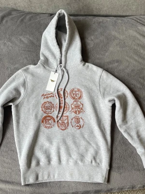 Grå hoodie från Maison Kitsuné - Snygg grå hoodie från Maison Kitsuné med tryck i rött på bröstet som visar olika emblem och loggor. Klassisk passform med huva och dragsko, ribbade muddar och mjukt material. Perfekt för en chill och trendig look.