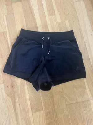Svarta velourshorts med snörning - Svarta shorts i mjuk velour från Juicy Couture med snörning i midjan och två bakfickor med knappar. Klassisk logga på ena benet och bekväm passform. Perfekta för en avslappnad och trendig look.
