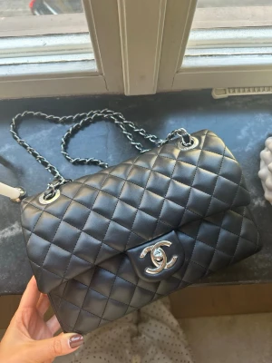 Svart quiltad väska  - Svart väska i quiltad veganläder med silverfärgad låsning och kedjerem. Klassisk rektangulär form som går att bära crossbody men även över axeln. Inget kvitto finns pga present, men i fint skick förutom en liten skråma i vänstra hörnet. Se bild, men inte ngt man tänker på! 