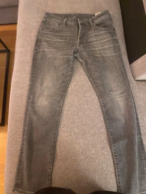 Grå tvättade jeans från J&J - Säljer ett par grå tvättade jeans från Nudie med klassisk femficksdesign och raka ben. Jeansen har snygga slitningar och kontrastsömmar, samt Nudie-logga på bakfickan. Perfekta för en avslappnad och trendig look.
