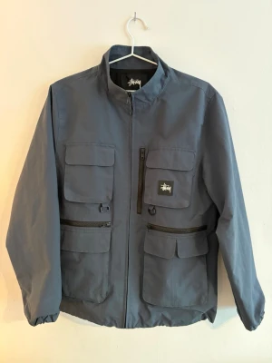 Blå vindjacka från Stussy - Snygg blå vindjacka från Stussy med fyra stora fickor framtill, dragkedja och hög krage. Jackan har en minimalistisk design med Stussy-logga på ena bröstfickan och elastiska ärmslut.