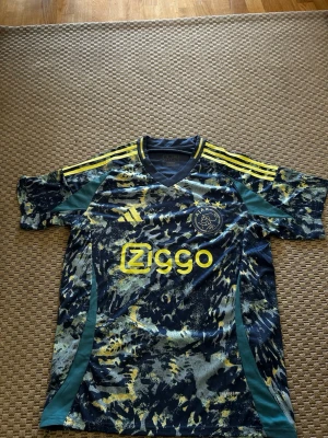 Ajax Amsterdam Jersey - Ajax Amsterdam matchtröja från Adidas med camo-mönster i mörkblått, gult och grönt. Tröjan har gula Adidas-ränder på axlarna, klubbmärke och sponsortryck på bröstet samt V-ringning. Perfekt för fotbollsträning eller att bära på läktaren.