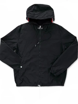 Svart Moncler vindjacka med huva - Stilren svart vindjacka med dragkedja framtill och justerbar huva. Jackan har en vit och röd rand på huvan baktill, elastiska muddar vid ärmsluten och en ficka med knapp. Perfekt för blåsiga dagar och enkel att matcha.