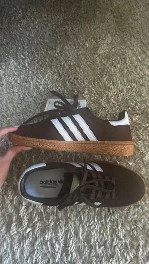 Bruna Adidas sneakers med vita ränder - Snygga bruna Adidas sneakers i mocka med klassiska vita ränder på sidorna och vit häl. Skorna har en låg profil och gummisula i brunt. Perfekta för dig som gillar retrostil och streetwear. Skön passform och tidlös design. Helt nya skicka prisförslag 