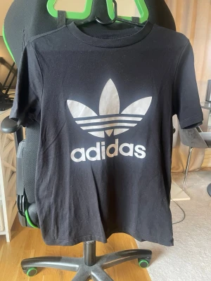 Svart Adidas t-shirt med logga - Svart t-shirt från Adidas med stor vit Trefoil-logga och text på bröstet. Klassisk rund halsringning och korta ärmar. Tillverkad i mjuk bomull som är skön att bära. Perfekt för en sportig och avslappnad stil.