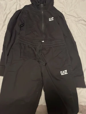 Svart EA7 loungewear set  - Svart loungewear set från EA7 Emporio Armani med dragkedja och huva på tröjan samt matchande byxor med dragsko i midjan. Båda delarna har EA7-logga tryckt i vitt. Perfekt för chill dagar hemma eller när du vill vara bekväm men ändå snygg. Skriv vid minsta tankar och funderingar 💭🤔Kan även komma med förslag på pris