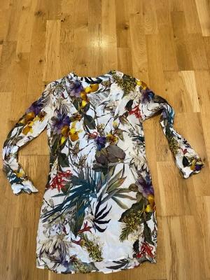 Zara Blommig långärmad blus stl M - Zara Snygg kort blus med färgstarkt blommönster i lila, gul, röd och gröna toner på vit botten. Långärmad modell med lätt v-ringning och flowy passform. Perfekt för dig som gillar att sticka ut med färg och mönster.