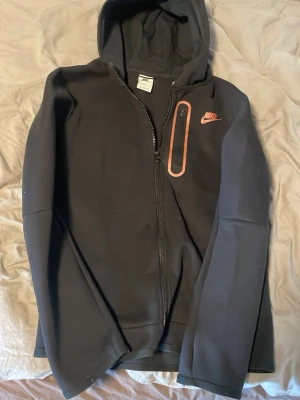 Svart Nike hoodie med dragkedja - Svart hoodie från Nike med dragkedja framtill och huva. Har en röd Nike-logga på bröstet och en snygg detalj med röd dragkedja på bröstfickan. Tillverkad i ett mjukt och stretchigt material, perfekt för en sportig och avslappnad stil. Skriv vid minsta tankar och funderingar 💭🤔 Kan även ge förslag på pris och komma överens 