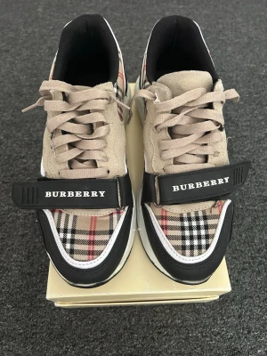 Burberry sneakers med klassiskt rutmönster - Snygga sneakers från Burberry med det ikoniska beige, svarta och röda rutmönstret. Skorna har beige mockadetaljer, svarta och vita inslag samt en bred svart rem med Burberry-logga över foten. Perfekta för dig som vill sticka ut med stil.