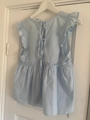 Ljusblå volangblus Zara - Supersöt ljusblå blus med volangdetaljer vid ärmhålen och v-ringning framtill. Blusen har knytning i ryggen och är tillverkad i ett mjukt bomullstyg. Perfekt för en fräsch och somrig look. Toppen är från Zara, står storlek M, men tycker definitivt mer än S