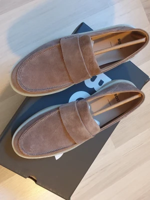 Bruna mocka loafers från BOSS - Snygga loafers i brun mocka från BOSS med klassisk penny strap och diskret logga på sidan. Skorna har en stilren design med rundad tå och ljus gummisula som ger en modern touch. Perfekta för dig som vill ha en clean och trendig look.