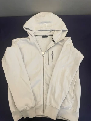 Vit hoodie från Sail Racing - Vit hoodie från Sail Racing med dragkedja och broderad logga på bröstet. Utrustad med huva och mjukt, fleecefodrat innerfoder för extra komfort. Perfekt för en chill och sportig stil. Klassisk passform och ribbade muddar.