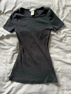 Svart ribbad topp från H&M - En svart, ribbad topp från H&M med rund halsringning och korta ärmar. Modellen är figurnära och enkel, perfekt att styla med jeans eller kjol 🩷 Tillverkad i mjukt material som känns skönt mot huden.