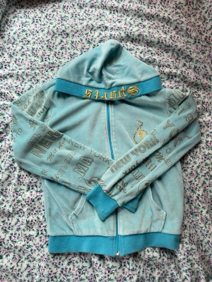 Turkos zipup hoodie med New York-tryck - Turkos hoodie från Afrodite med dragkedja, huva och ribbade muddar. Ärmarna har gråa New York-inspirerade tryck och på bröstet finns en guldfärgad brodyr. Materialet är mjukt och känns som velour. Perfekt för en chill och avslappnad stil. Det står S på lappen men passar som en XS