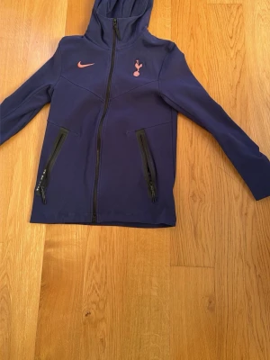 Mörkblå Nike Tottenham hoodie - Snygg mörkblå hoodie från Nike med Tottenham Hotspur-logga på bröstet och orange Nike-swoosh. Dragkedja framtill och två fickor med svarta dragkedjor. Perfekt för dig som gillar fotboll och vill ha en sportig stil. Använt ett par gånger, men är som helt ny skick