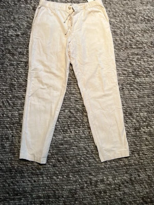 Beiga linnebyxor med dragsko - Snygga beiga linnebyxor med avslappnad passform och dragsko i midjan. Byxorna har resår i midjan, knäppning framtill och två bakfickor med knapp. Perfekta för en chill och stilren look.