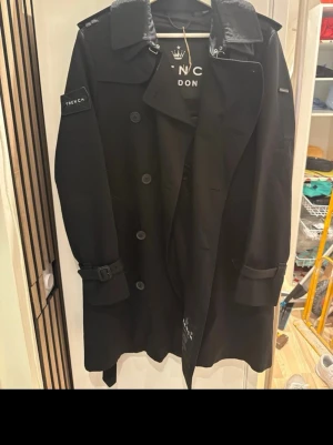Svart kappa från  trench London - Stilren svart trenchcoat från NYC London med klassisk dubbelknäppning, bälte vid ärmslut och axelklaffar. Jackan har detaljer som 'TRENCH' patch på ärmen och bred krage. Perfekt för dig som vill ha en tidlös och trendig look. Helt ny och fin köpt för 5500kr 